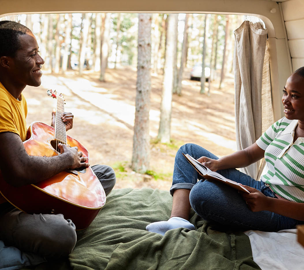 African young couple resting in van, playing guitar and reading book, camping in forest