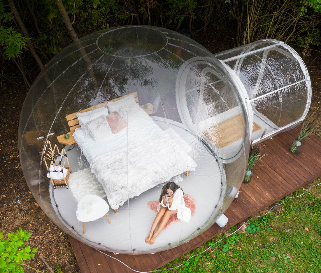 Transparent bubble tent at glamping, Lush forest around and interior, Woman with a cup looking outside, drone view